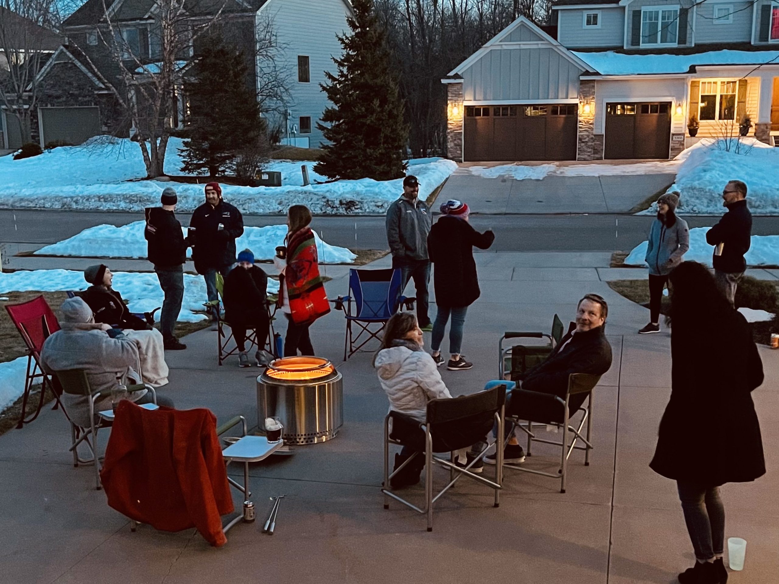 Several people are gathering around a fire pit in a dirveway. There is snow on the ground, and it's a fun cheerful scene.