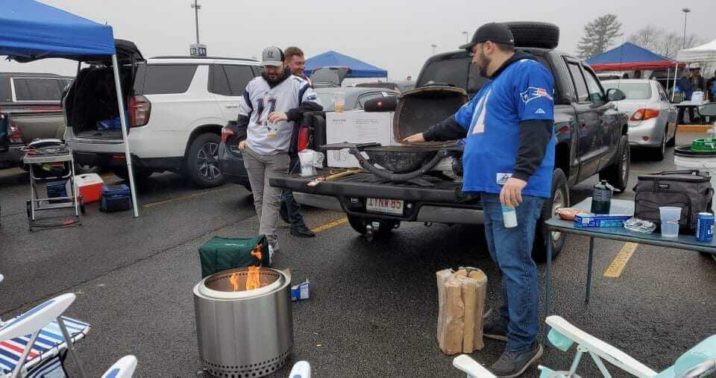 Tailgate goers celebrate a win around their fire pit. It's sitting on the ground next to a truck bed with an open tailgate, most likely in a whole lot full of other tailgaters.
